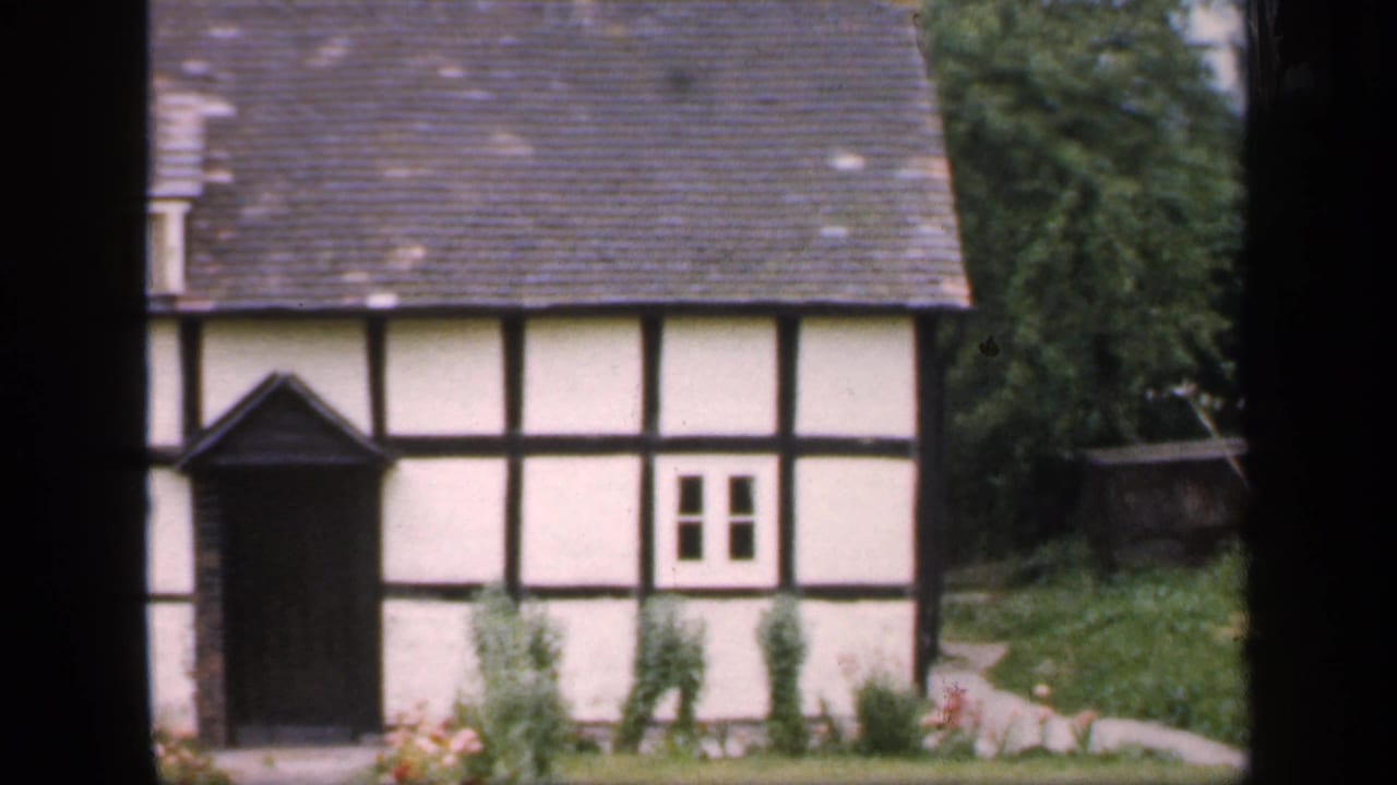 Half-timbered cottage exterior with tiled roof, small window, dark entrance, and garden greenery.