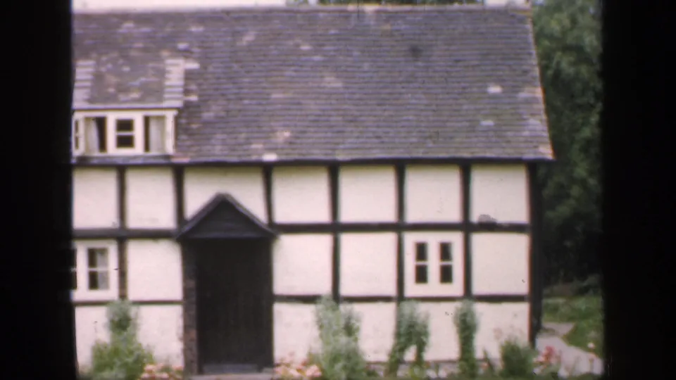 1949 England home-movie view of the cottage front, showing timber framing, a dormer, dark porch, small windows, and garden plantings.