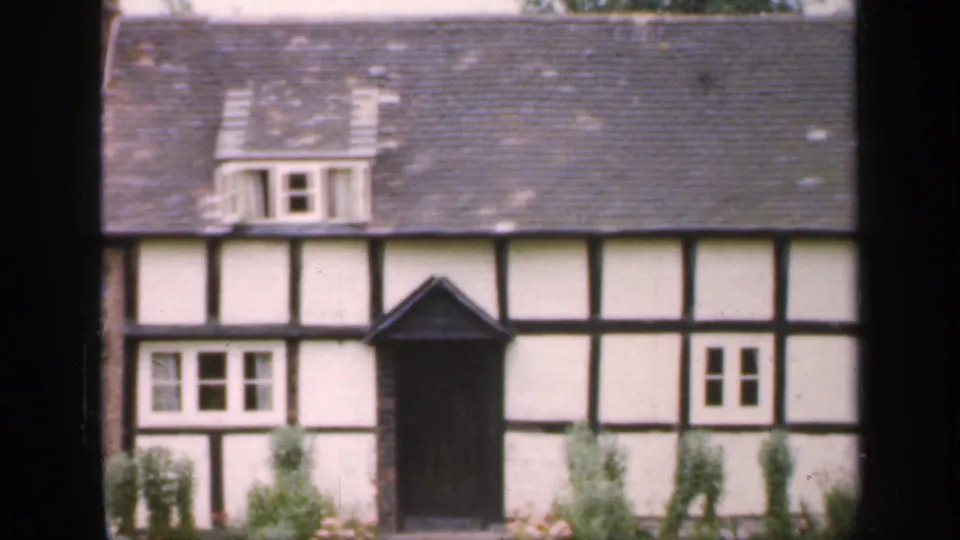 1949 England frame showing the cottage facade with white plaster panels, dark timber beams, a central porch, dormer, windows, and greenery.