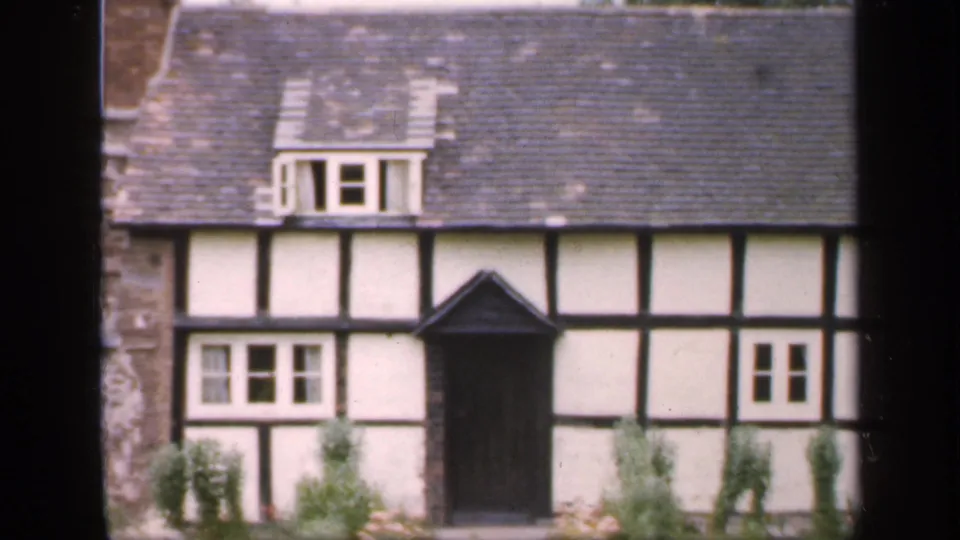 1949 England home-movie frame showing a Tudor-style house with dark timber framing, white wall panels, slate roof, chimney, and shrubs.