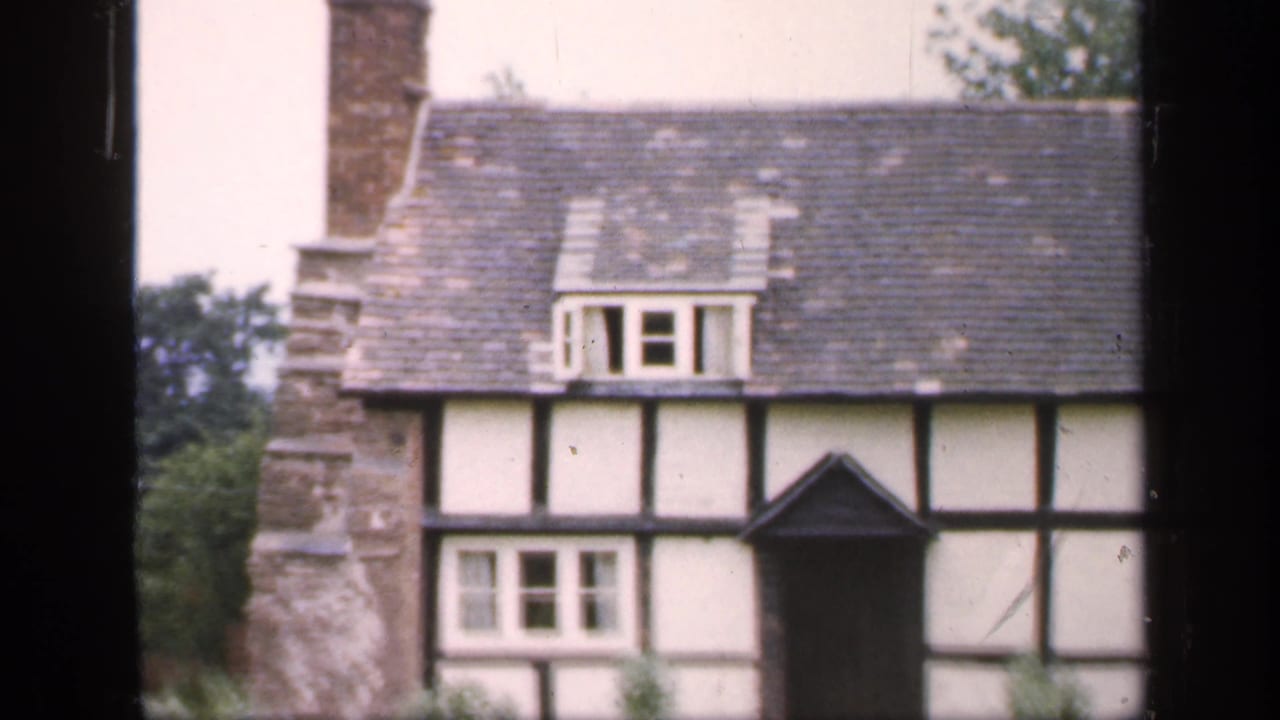 1949 England frame of a Tudor-style house facade, with a prominent chimney, slate roof, dormer window, dark doorway, and garden foreground.