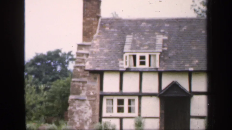 1949 England home-movie frame with the house partly cropped, showing chimney masonry, slate roof, dormer window, dark doorway, and trees behind.
