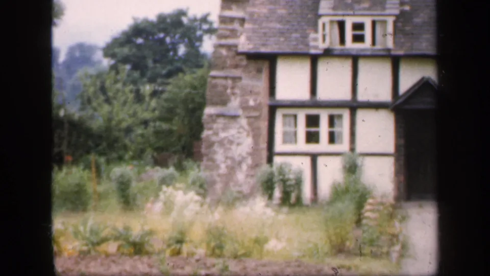 1949 England frame showing the right side of a Tudor-style house, with timber framing, chimney, dormer window, dark doorway, and overgrown garden.