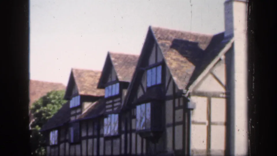 1949 England home-movie view of gabled half-timbered houses, dark beams, tiled roofs, foliage, and a pale chimney near the right edge.