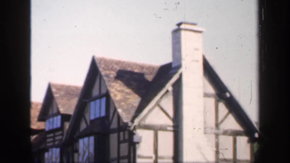 1949 England frame with Tudor-style houses, black timber detailing, steep tiled roofs, a prominent chimney, and dark foreground masking.