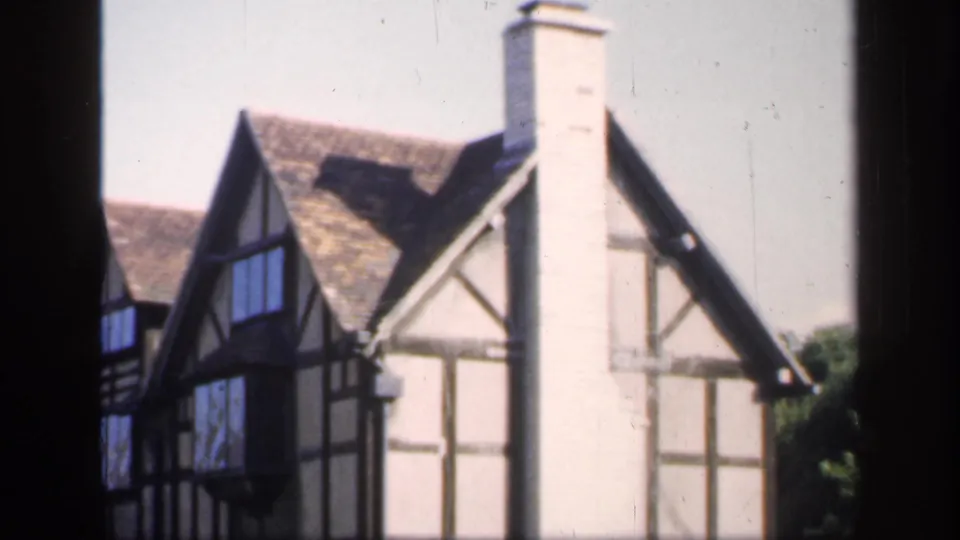 1949 England home-movie frame showing timber-framed gables, pale plaster walls, a tall chimney, and shrubs near the building edge.