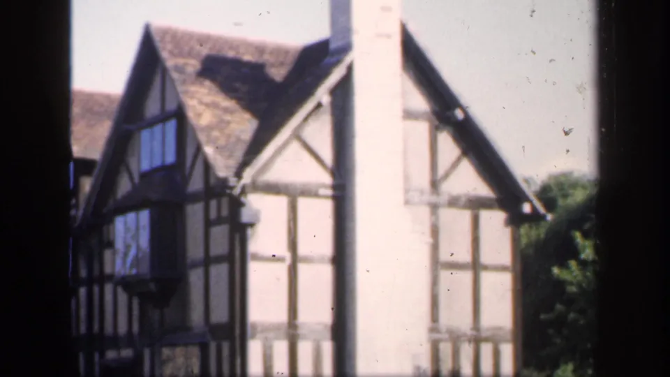 1949 England frame showing a Tudor-style residential exterior with black timber framing, steep roof planes, a tall chimney, and side greenery.