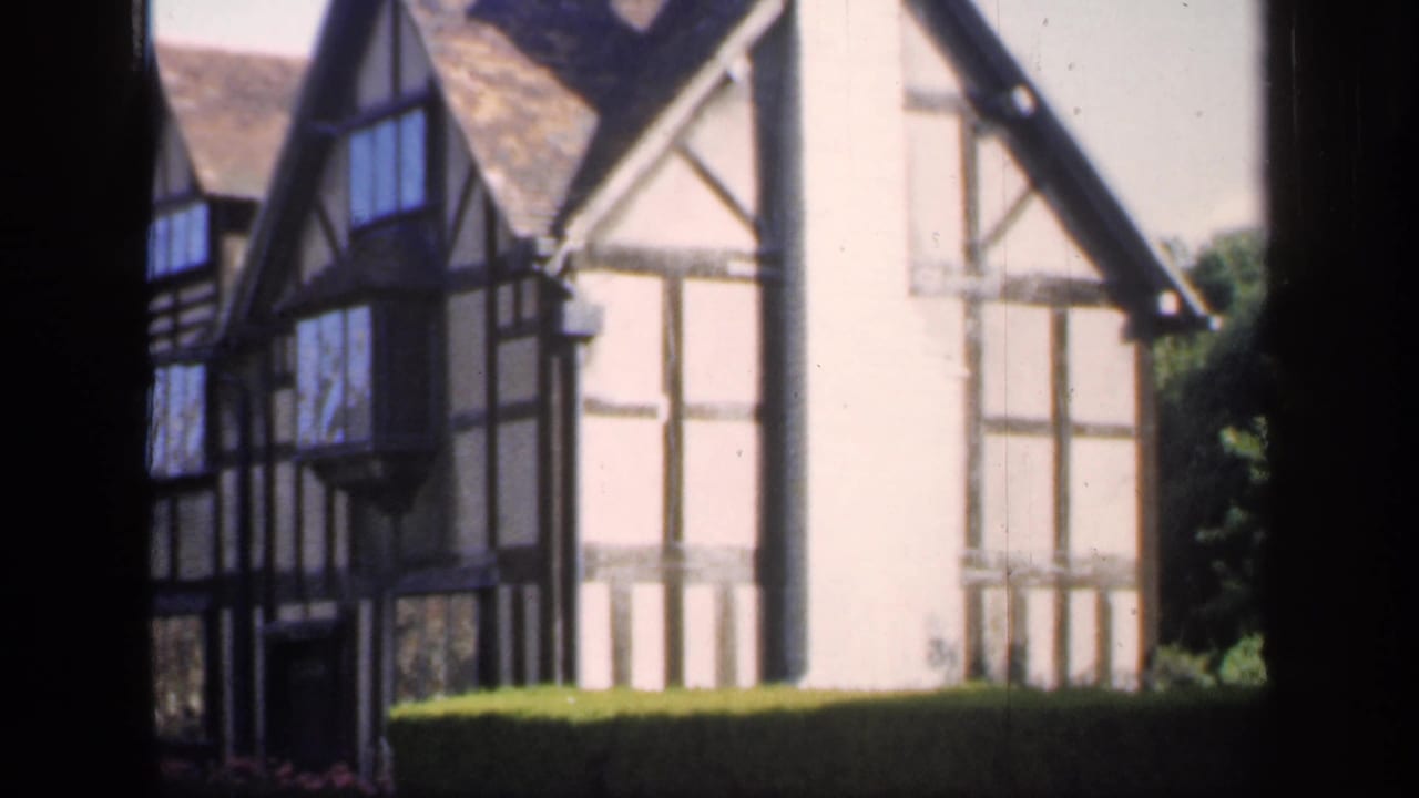 The camera holds on an English timber-framed house with gables, white wall panels, and a hedge in the foreground.