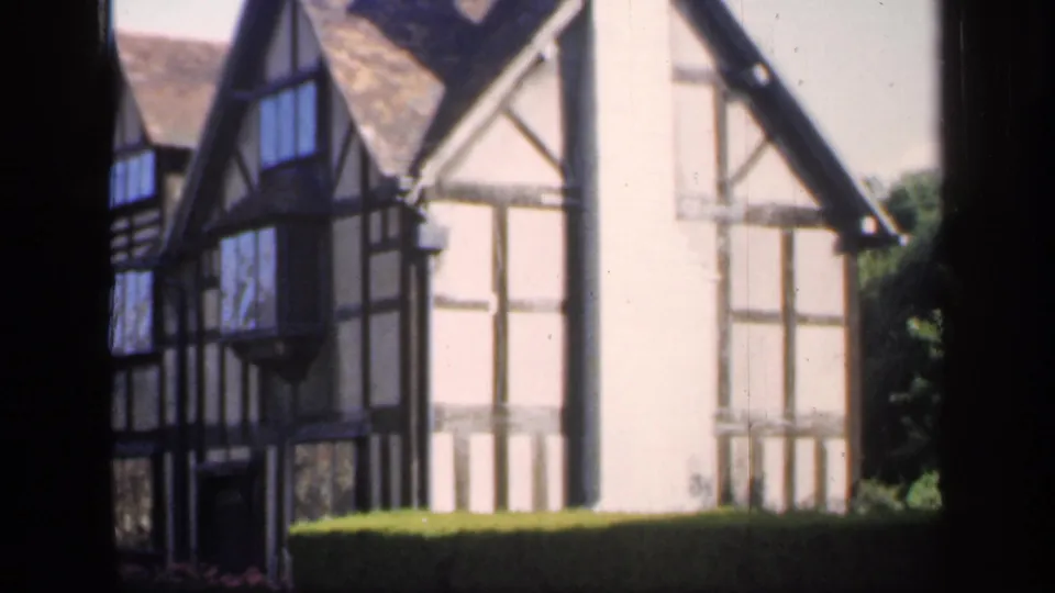 The camera holds on an English timber-framed house with gables, white wall panels, and a hedge in the foreground.