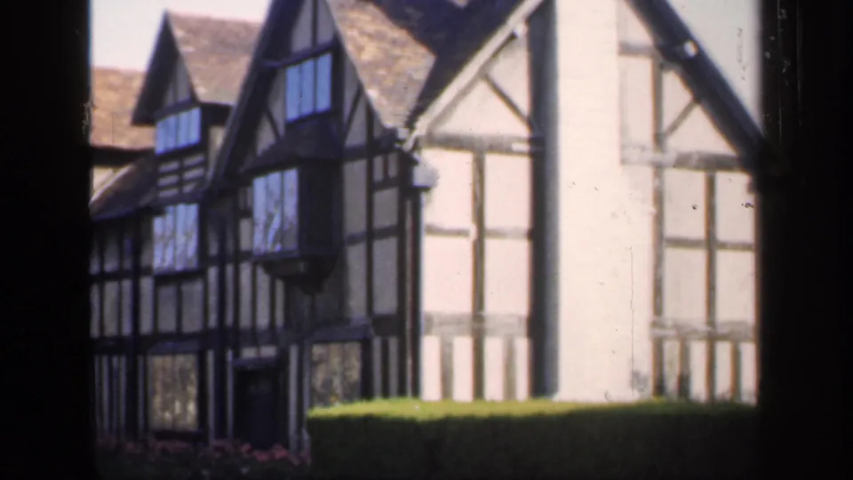 1949 home-movie frame of a traditional English timber-framed facade, with dark beams, windows, rooflines, and a trimmed hedge.