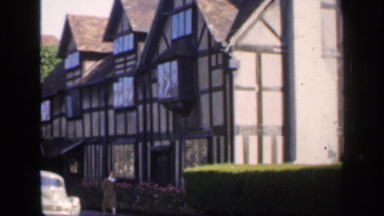 1949 England 8mm frame showing a Tudor-style house frontage, trimmed hedge, pale vintage car, and a person near the building.