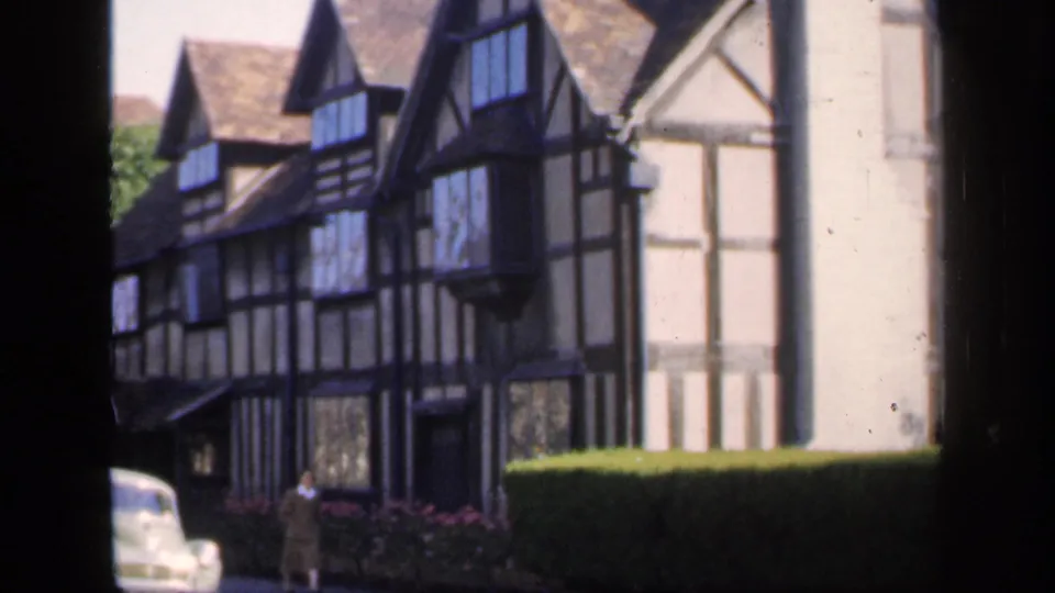 1949 England 8mm frame showing a Tudor-style house frontage, trimmed hedge, pale vintage car, and a person near the building.