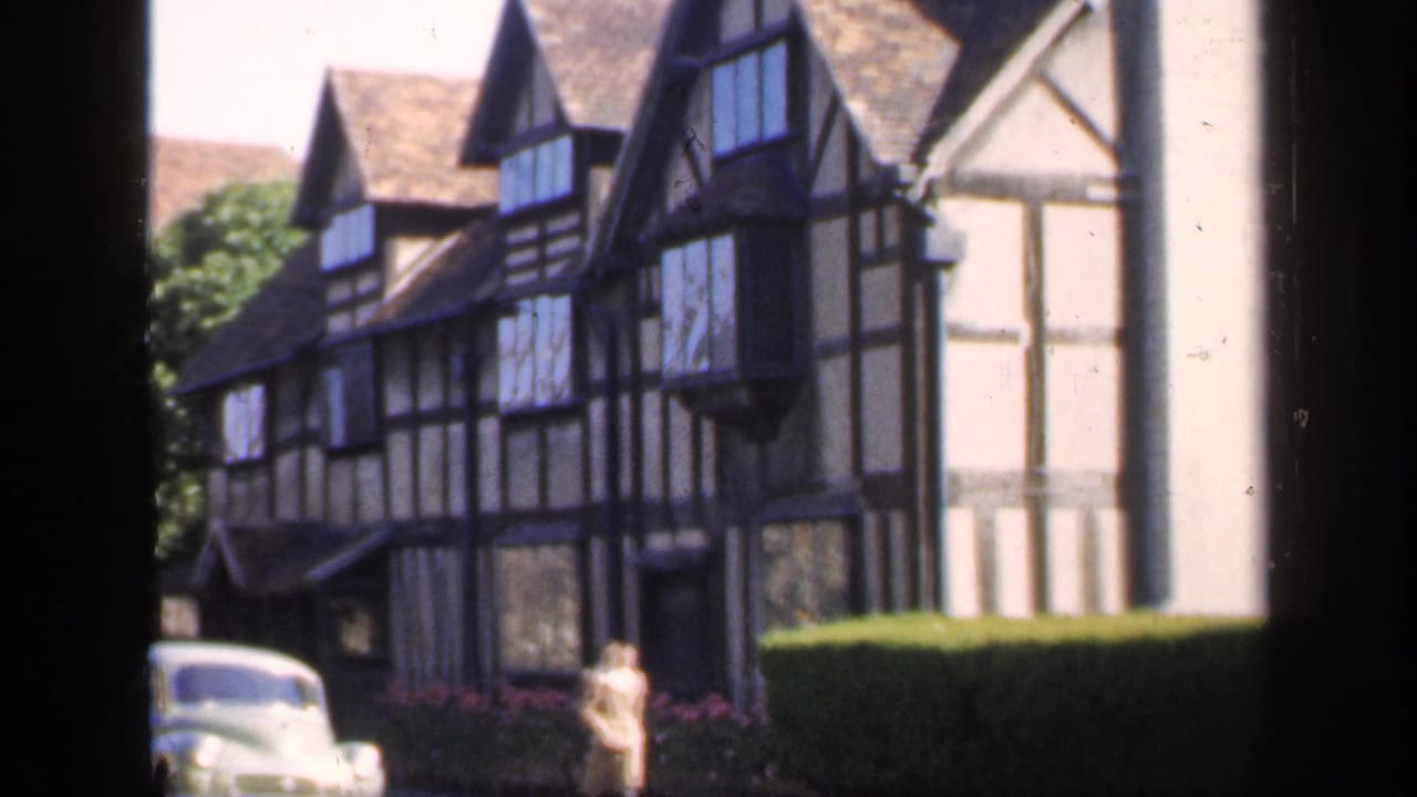 1949 England 8mm frame of a woman walking past a Tudor-style house, clipped hedge, and pale parked car.