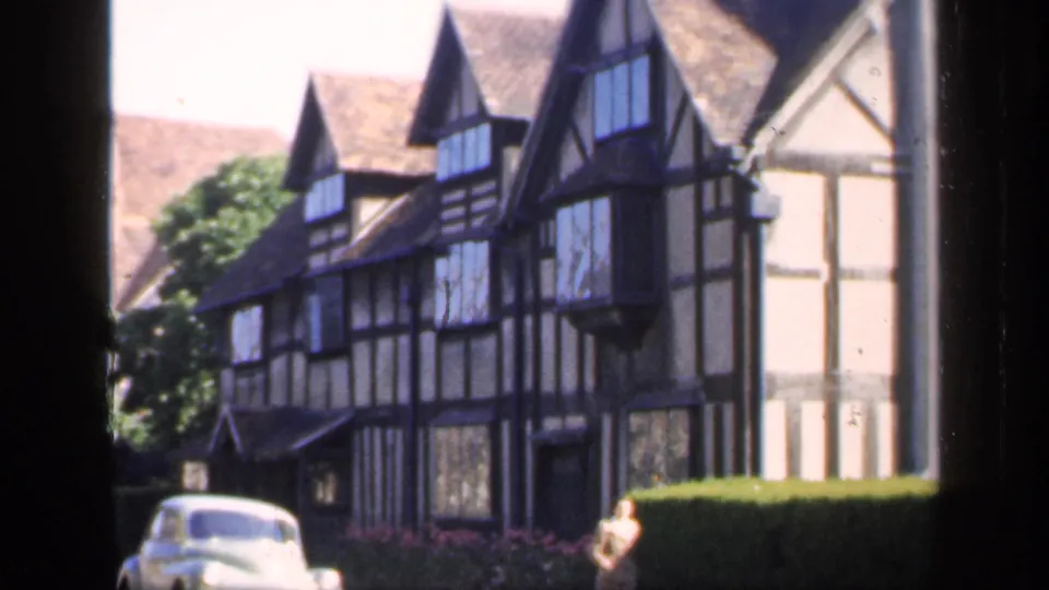 1949 England 8mm frame with a closer view of Tudor-style timber framing, hedge, pale car, and a woman at the frontage.