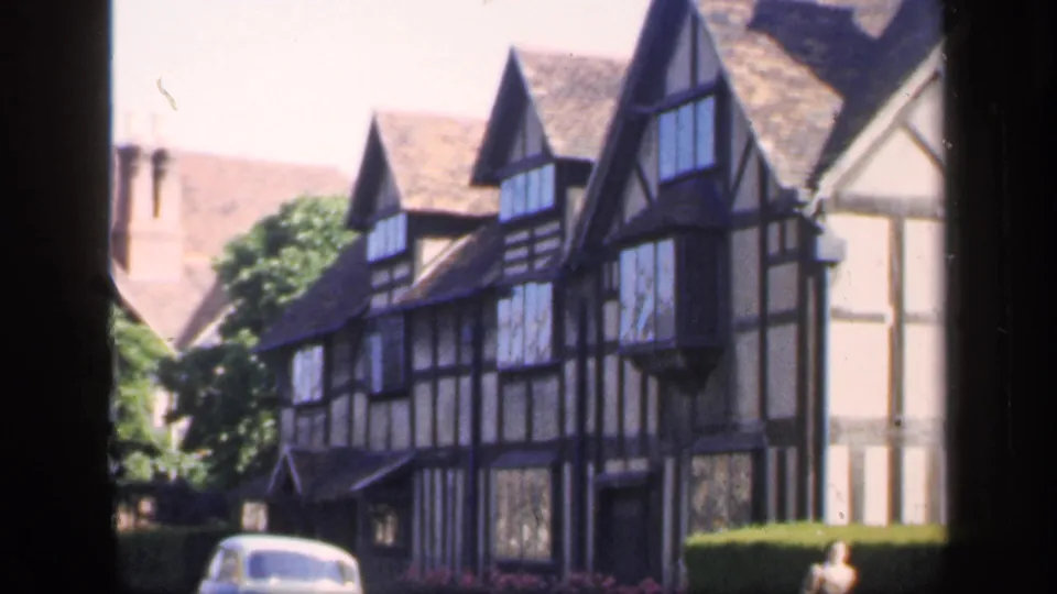 1949 England 8mm frame showing the Tudor-style building, clipped hedge, pale vintage car, and woman near the right side.