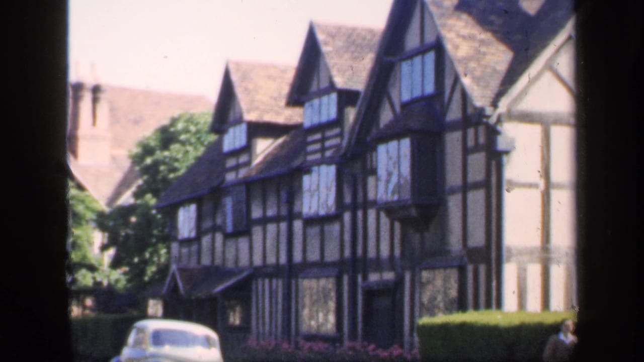 1949 England home-movie frame showing a half-timbered Tudor-style building, steep rooflines, hedges, and a light-colored car at street level.