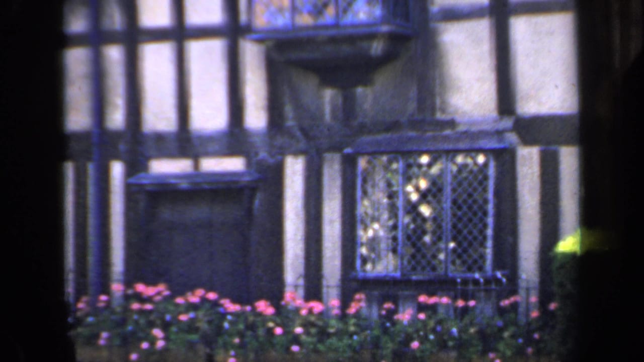 Closer view of half-timbered walls, leaded windows, a small projecting balcony, and pink flowers.