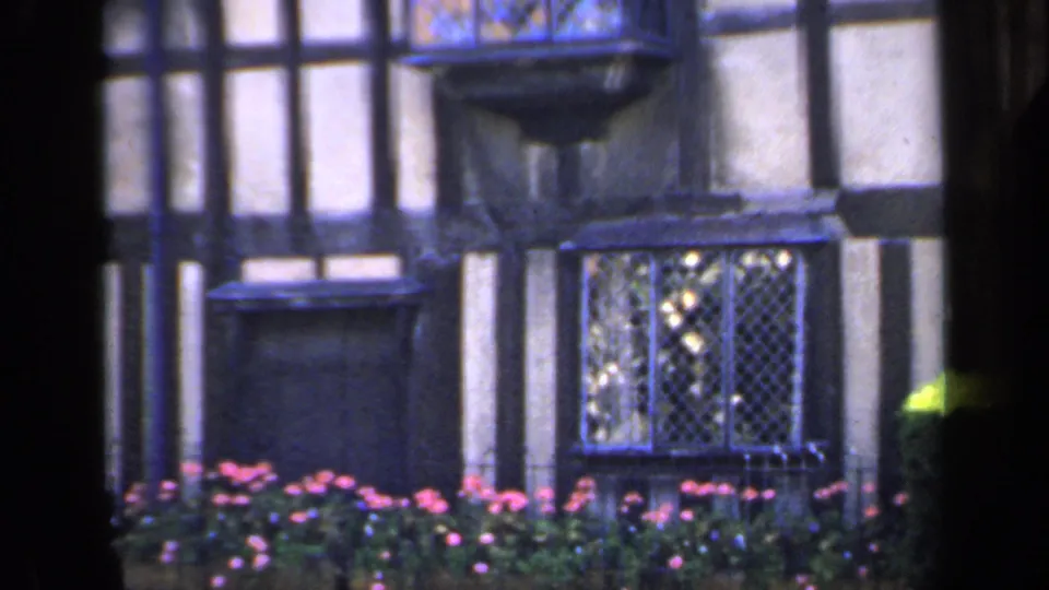 1949 England frame with the camera closer to the Tudor-style facade, showing leaded windows, dark timbering, and a flower bed below.