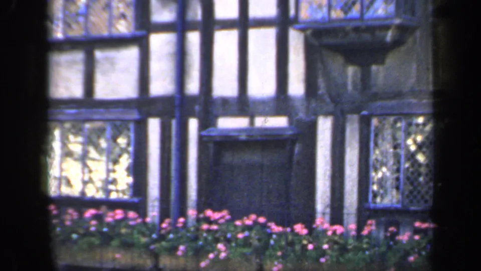 1949 England home-movie detail of the half-timbered facade with diamond-pane windows, projecting balcony feature, and garden flowers.