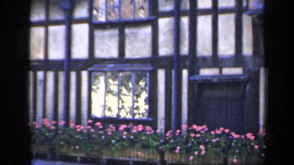 The pan ends on a 1949 England view of the Tudor-style wall, bright leaded window, flower bed, and dark vertical foreground edge.