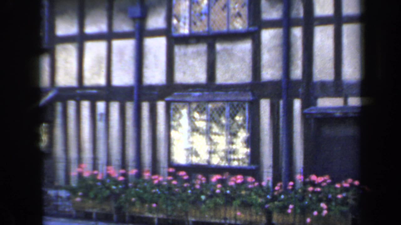 1949 England home-movie frame showing a Tudor-style house facade with dark timbering, pale panels, leaded windows and flower boxes.