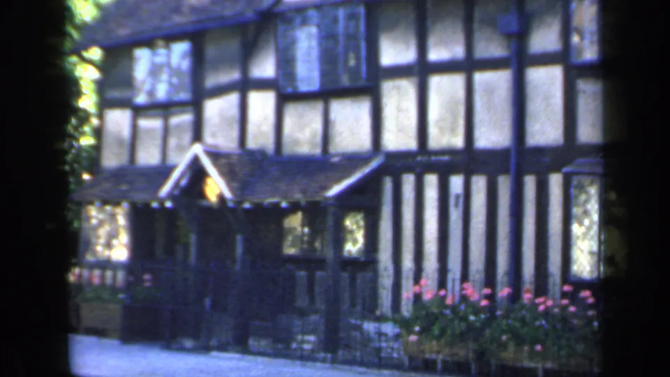 1949 England frame centering the gabled doorway of a Tudor-style house, with half-timbering, leaded windows and pink flowers nearby.