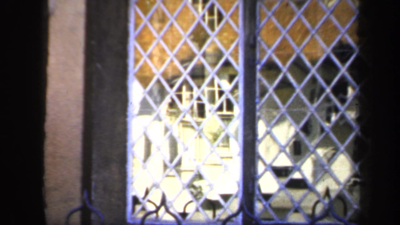 1949 England 8mm frame looking through a diamond-patterned grille toward brick walls and pale structures at a heritage site.