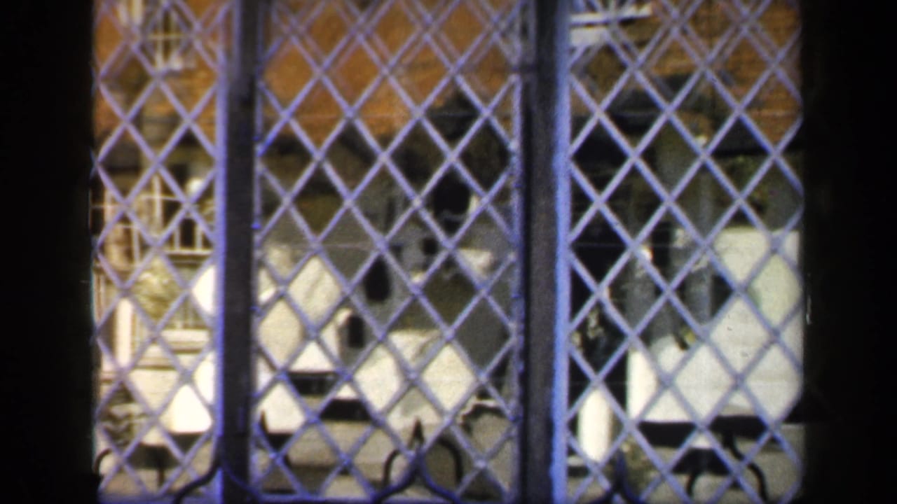 1949 England home-movie frame showing a leaded grille and dark timber division with brick and pale details beyond.
