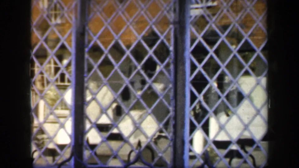 1949 England home-movie frame showing a leaded grille and dark timber division with brick and pale details beyond.