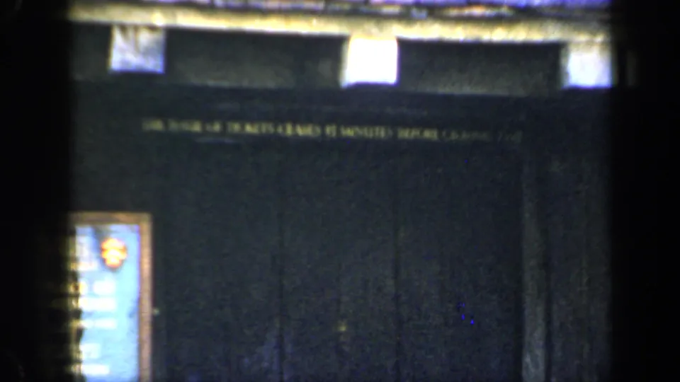 1949 England home-movie frame showing dark wall signage, an upper ticket-office style sign and a small framed plaque near the lower left.