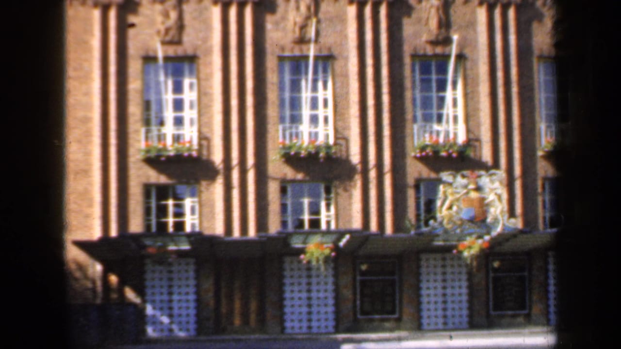 1949 England 8mm frame showing a brick building with vertical columns, multi-pane windows, flower boxes and a decorative crest above the entrance.