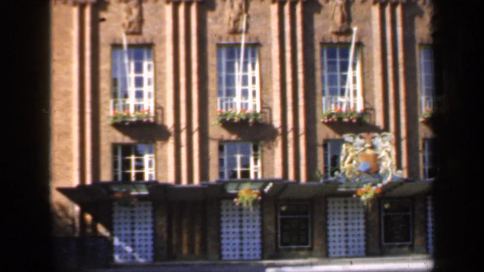 1949 England 8mm frame showing a brick building with vertical columns, multi-pane windows, flower boxes and a decorative crest above the entrance.