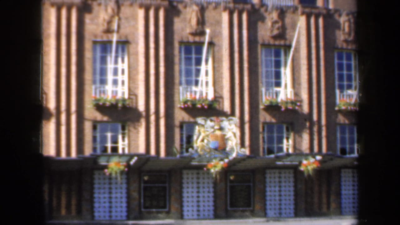 1949 England home-movie frame of a historic-looking brick facade, showing repeated windows, flower boxes and a coat of arms near the entrance.