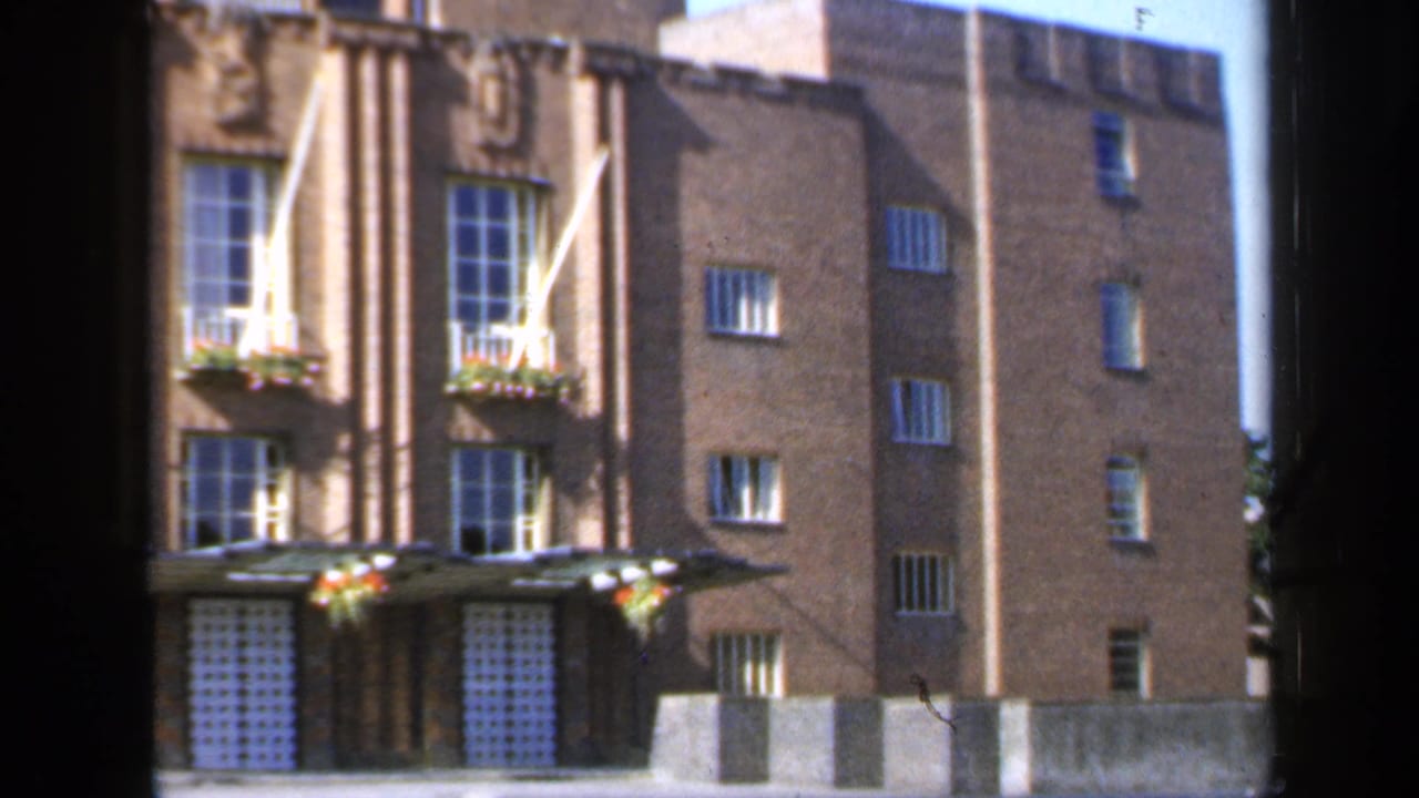 1949 England 8mm frame showing a formal brick building facade with vertical columns, tall windows, flower boxes, and white flagpoles.