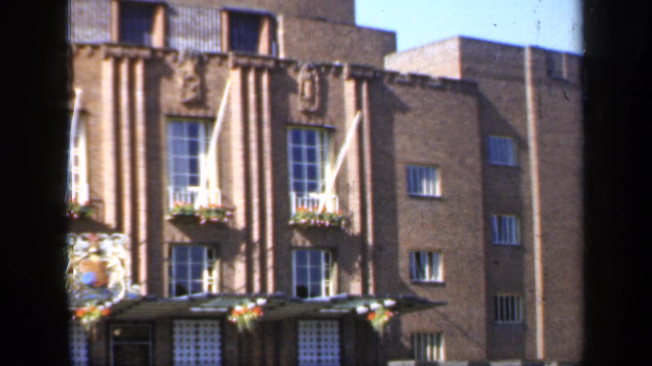 1949 England 8mm frame of a sunlit formal brick facade, showing white flagpoles, flower boxes, tall windows, and carved relief details.