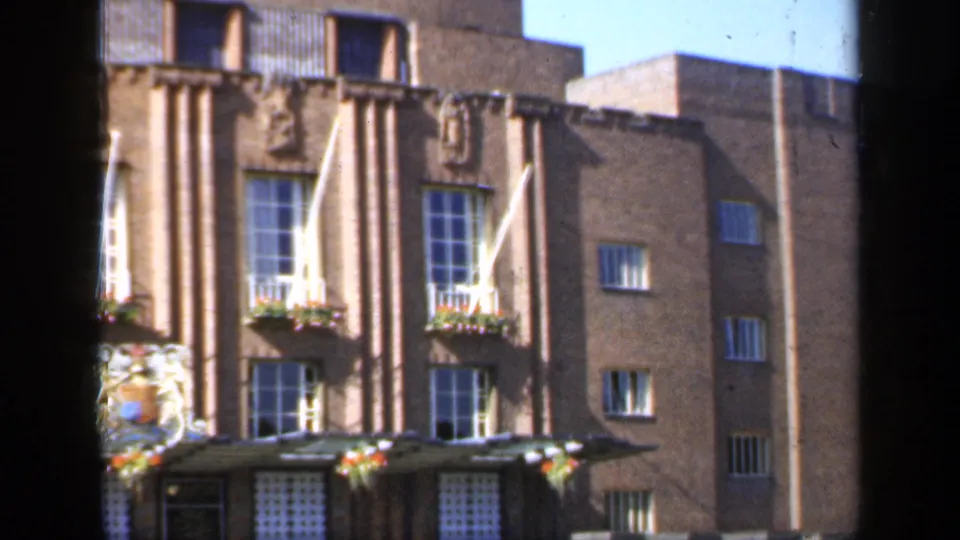 1949 England 8mm frame of a sunlit formal brick facade, showing white flagpoles, flower boxes, tall windows, and carved relief details.