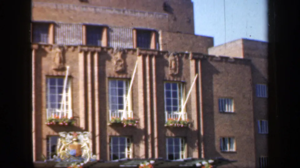 1949 England 8mm frame showing the building from a low angle, with brick walls, tall windows, white flags, flower boxes, and stone reliefs.