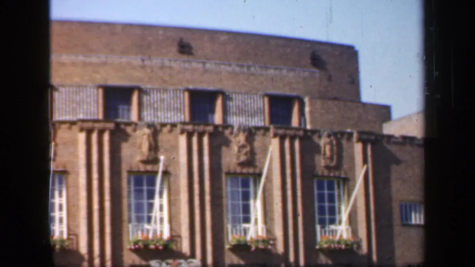 1949 England 8mm frame of the upper portion of a formal brick facade with repeated windows, white flagpoles, flower boxes, and roofline.