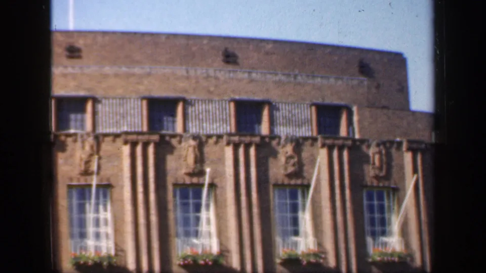 1949 England home-movie frame showing a brick Art Deco-style facade with sculptural figures, tall windows and dark window-frame borders.