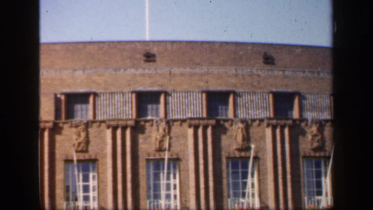 1949 England frame of a brick architectural facade with repeated windows, vertical columns and decorative relief figures under a pale sky.
