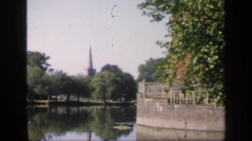 1949 England home-movie frame looking across a lake or canal toward trees, a stone embankment and a distant church spire.