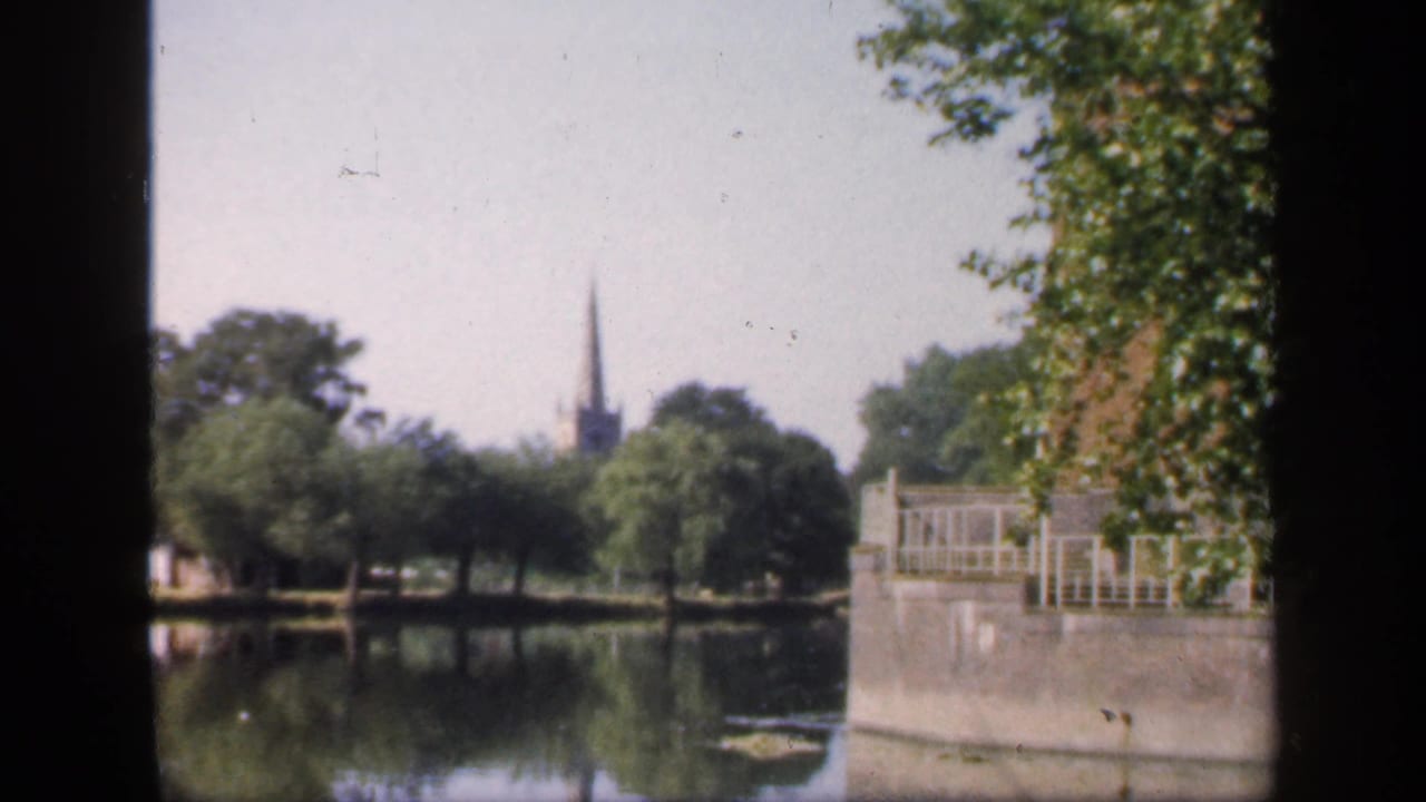 1949 England home-movie frame showing a still lake, leafy trees, waterside railings, and a distant church spire seen through dark framing.