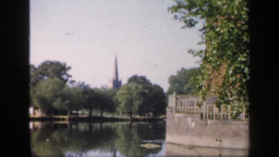 1949 England home-movie frame showing a still lake, leafy trees, waterside railings, and a distant church spire seen through dark framing.
