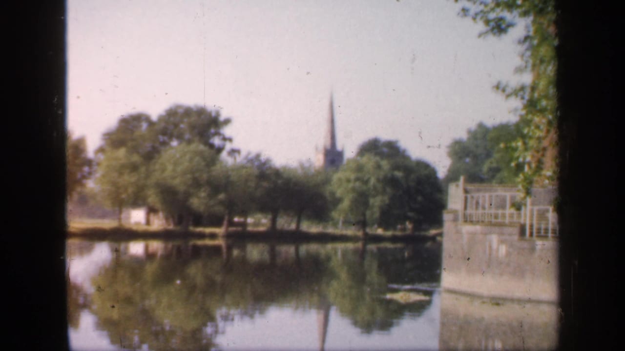 1949 England park view with still water, reflected trees, a distant church spire, and a waterside railing partly blocked by dark frame edges.