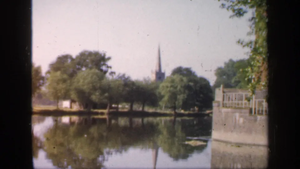 1949 England park view with still water, reflected trees, a distant church spire, and a waterside railing partly blocked by dark frame edges.