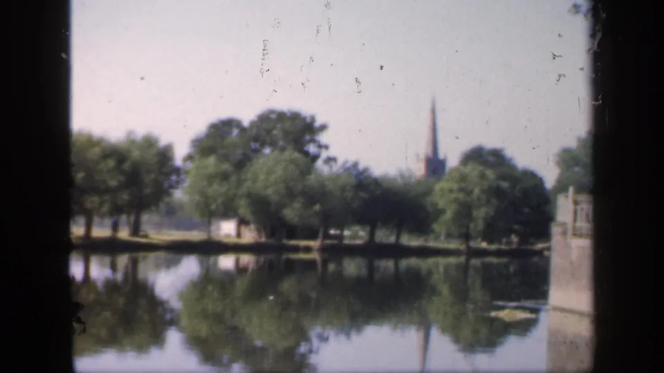 1949 England 8mm frame of a calm park lake, dense green trees, a small pale structure, and a distant church spire reflected in the water.