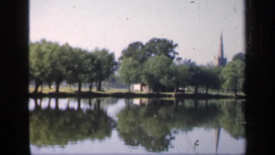 1949 England home-movie view of a quiet lake, reflected trees, a distant church spire, and a small pale structure along the opposite bank.