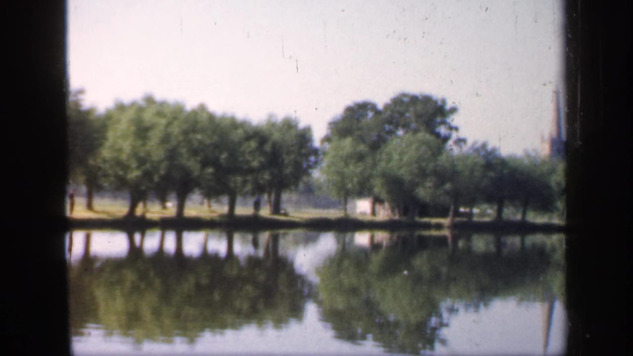 1949 England home-movie frame showing a calm river bordered by trees, with reflections on the water and a distant spire partly visible.