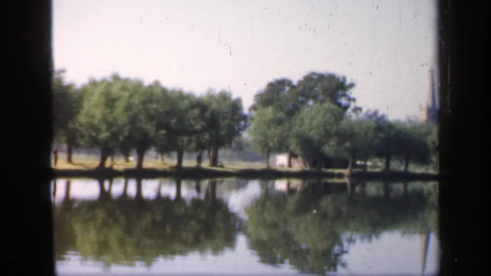 1949 England home-movie frame showing a calm river bordered by trees, with reflections on the water and a distant spire partly visible.