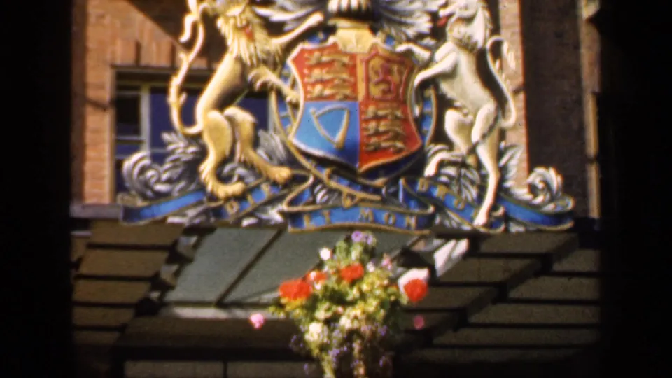 1949 England home-movie frame with a close view of the theatre crest, brick wall, canopy edge, and hanging basket of bright flowers.